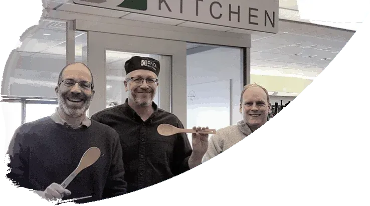 DataKitchen founders Chris Bergh, Eric Estabrooks and Gil Benghiat under the office KITCHEN sign, holding wooden spoons, one in a DataKitchen chef's hat